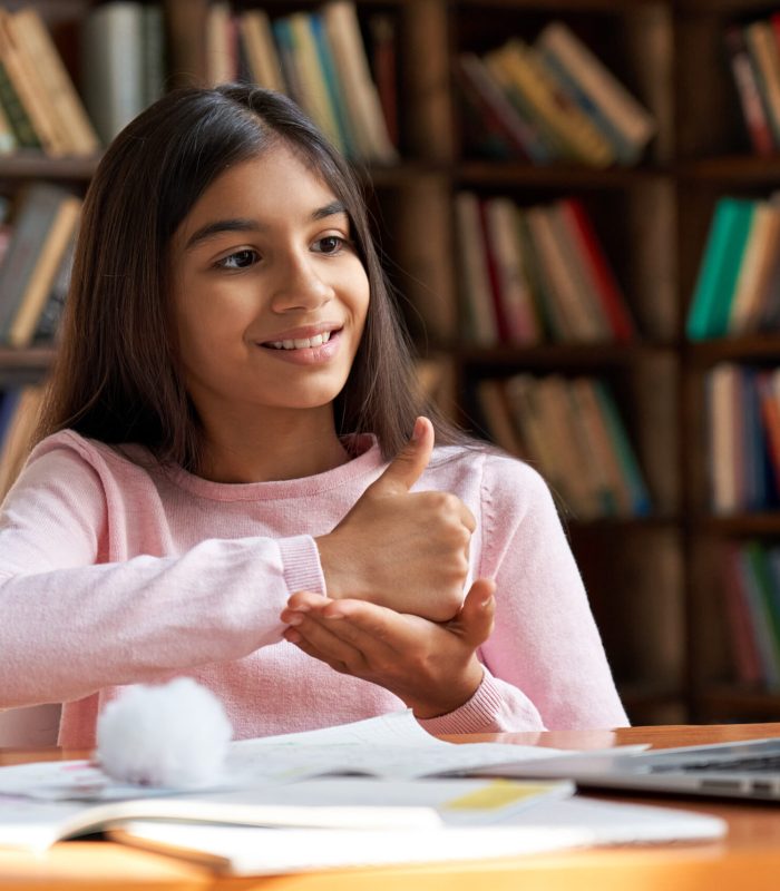 Smiling indian latin deaf disabled child school girl learning online class on laptop communicating with teacher by video conference call using sign language showing hand gesture during virtual lesson.